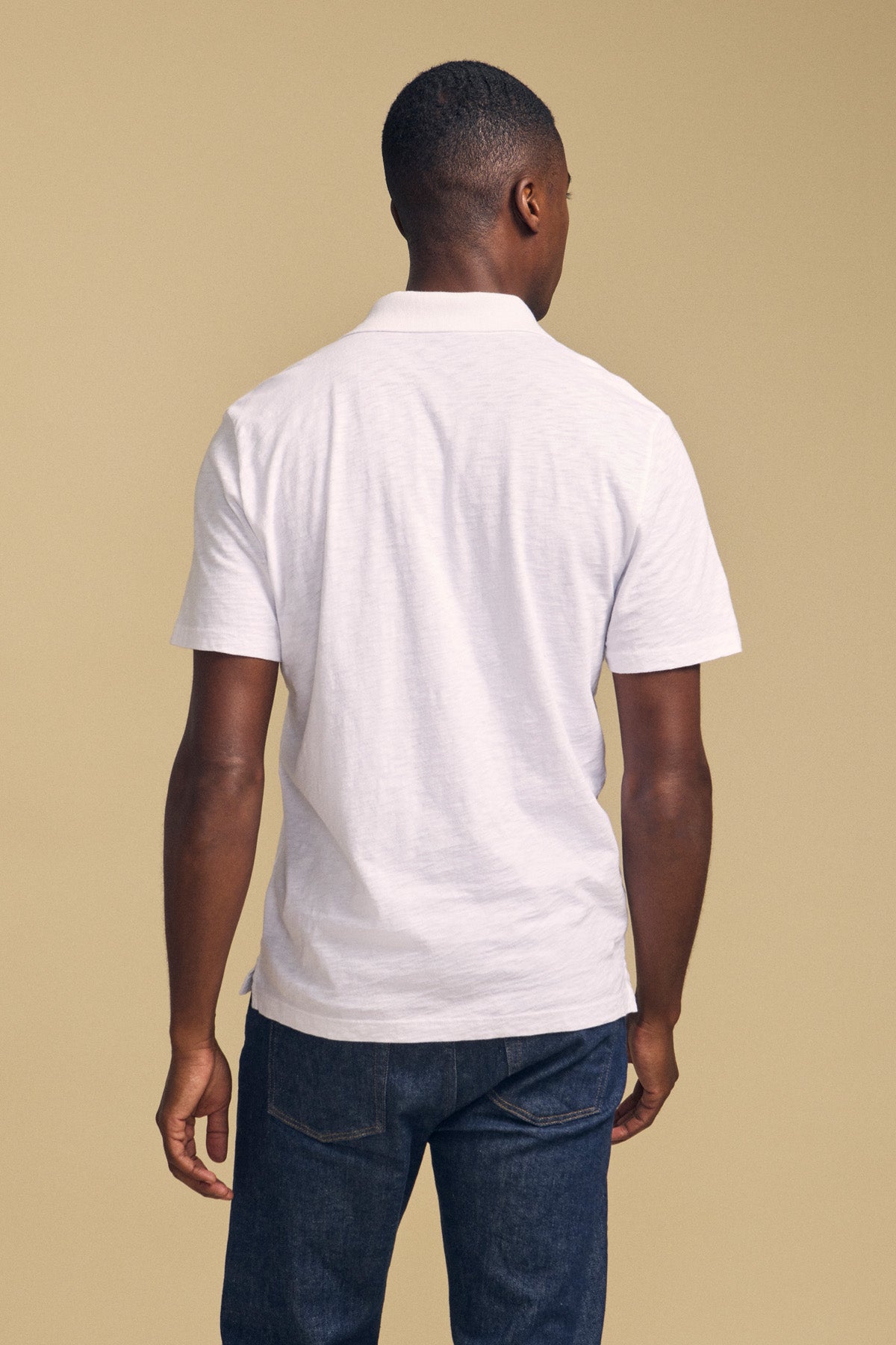 A man stands with his back to the camera, wearing the MORRIS POLO by Velvet by Graham & Spencer in white and dark blue jeans, set against a plain beige background.