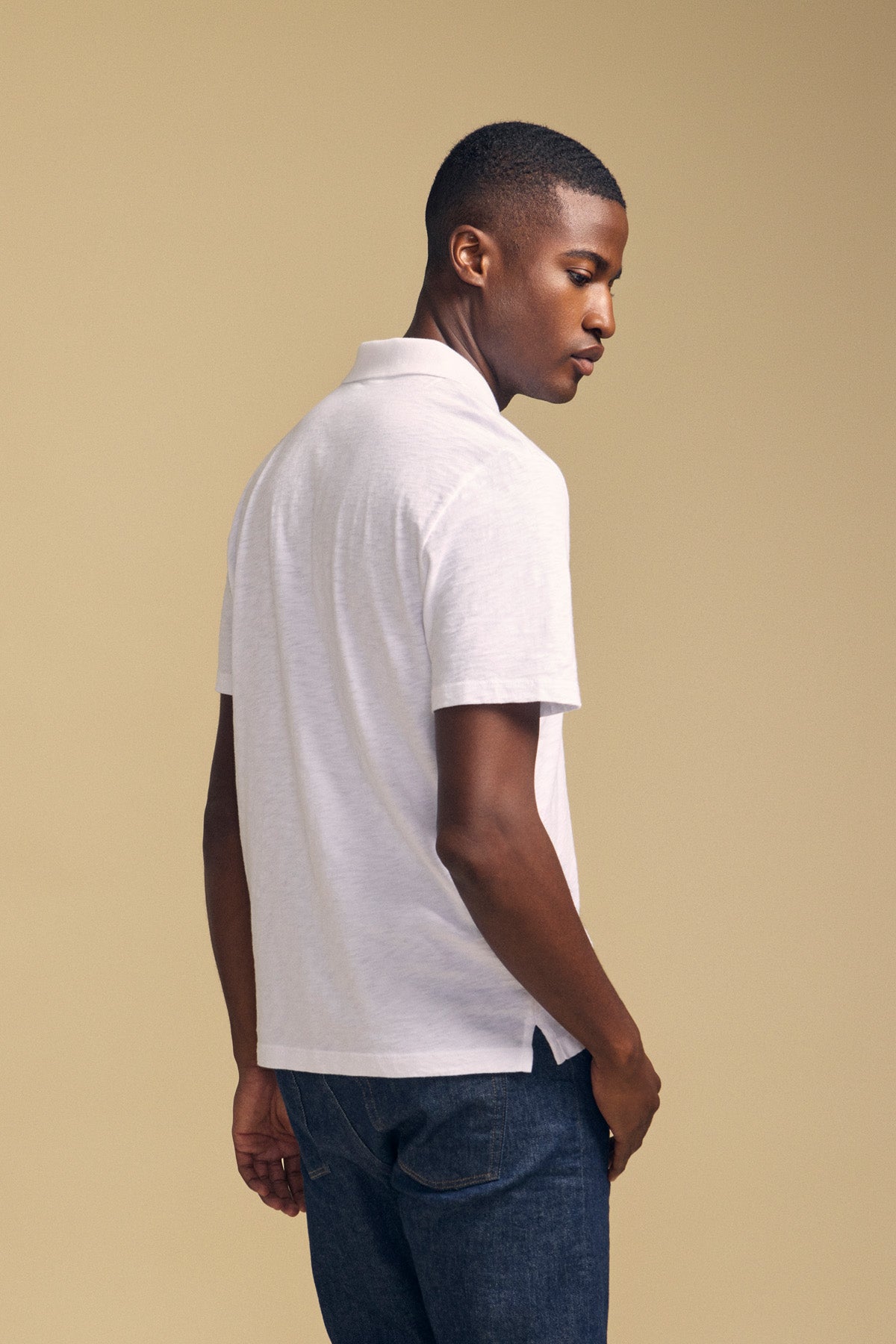 A person stands sideways against a plain beige background, wearing the MORRIS POLO by Velvet by Graham & Spencer— a white cotton slub short-sleeve polo—paired with dark blue jeans.