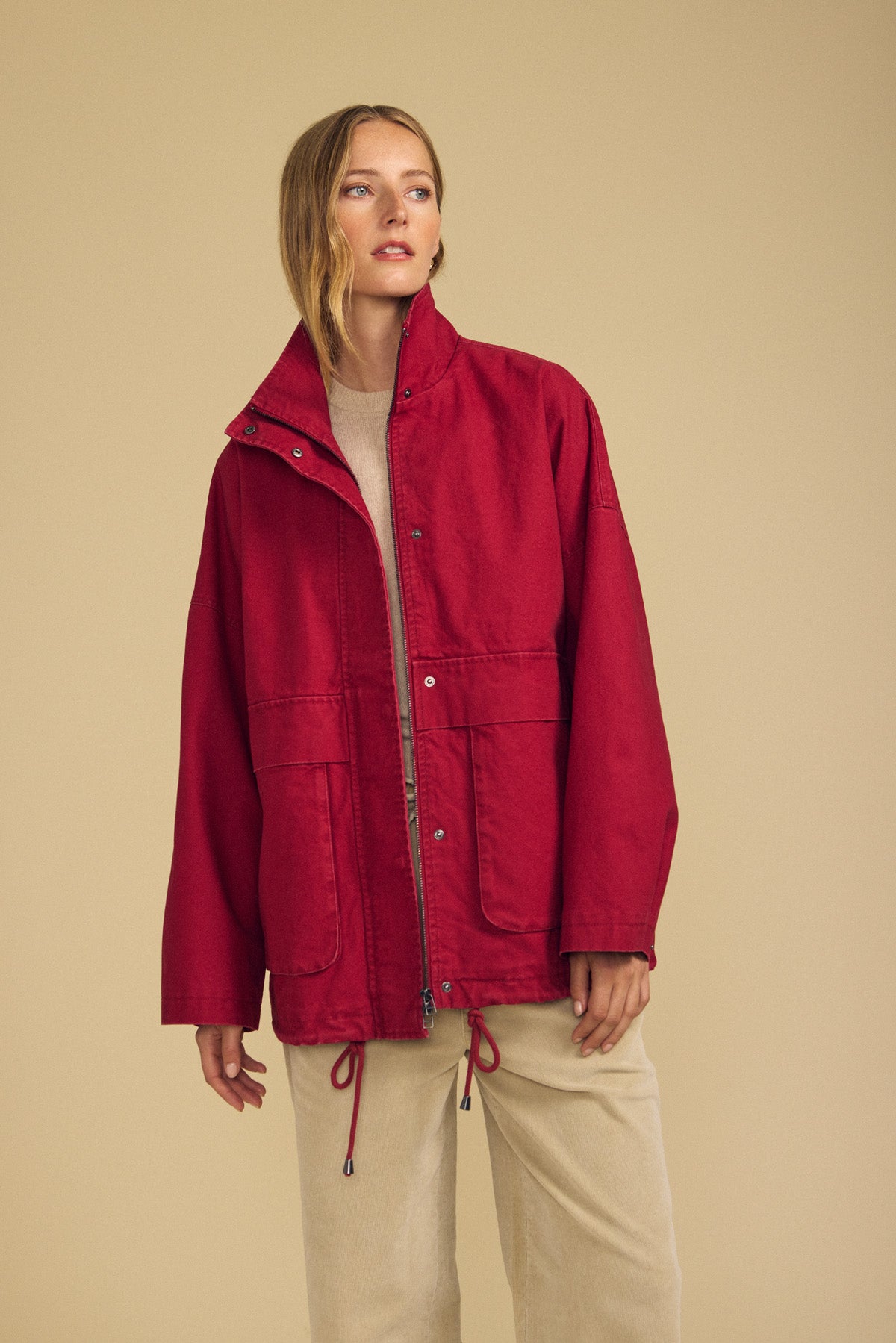 A person stands in a beige room wearing the HUNTER PARKA by Velvet by Graham & Spencer—a bright red cotton canvas piece featuring oversized front patch pockets—layered over a light beige top and pants, looking slightly to the side.