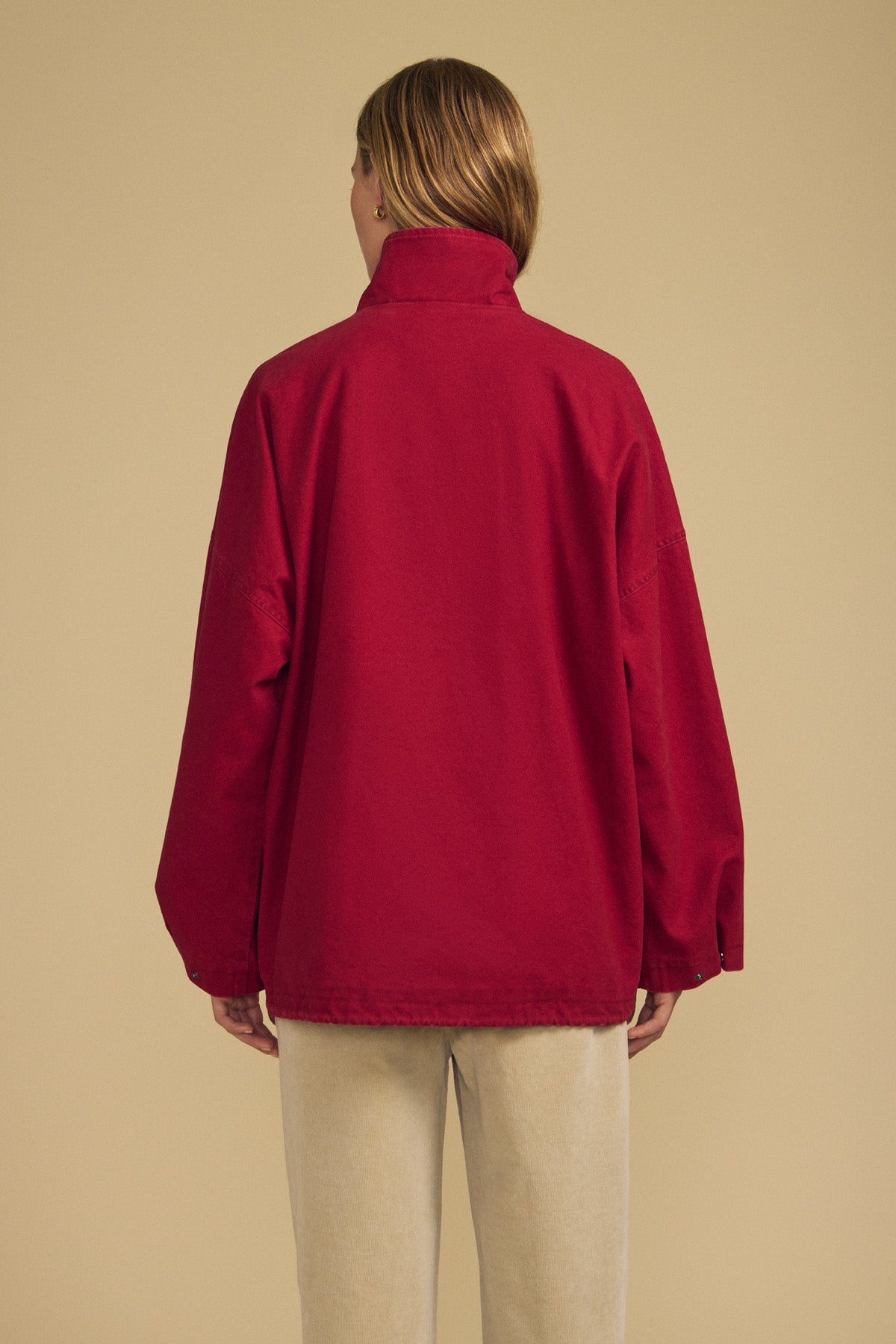 A person stands with their back to the camera, modeling the HUNTER PARKA in red cotton canvas by Velvet by Graham & Spencer, paired with beige pants against a plain tan background—an effortless display of versatile cold-weather style.