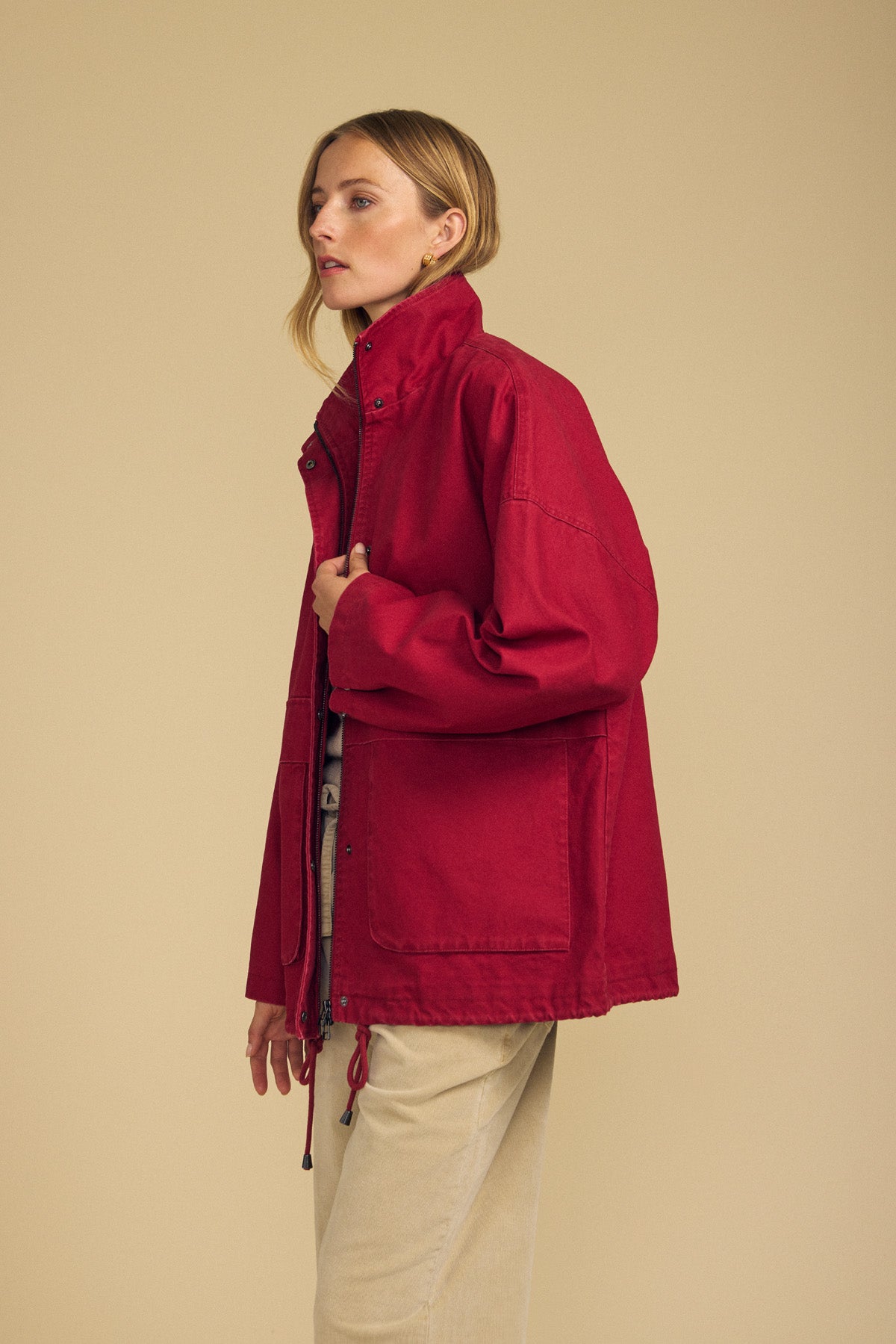A woman stands in profile against a plain beige background, wearing the HUNTER PARKA by Velvet by Graham & Spencer—a red cotton canvas design with oversized front patch pockets—layered over a beige outfit, making it an ideal choice for stylish cold-weather wear.