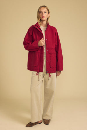 A person stands against a beige background wearing the HUNTER PARKA by Velvet by Graham & Spencer—a striking red cotton canvas jacket with oversized front patch pockets—layered over a beige top and wide-legged beige pants, finished with brown shoes for a perfect cold-weather ensemble.