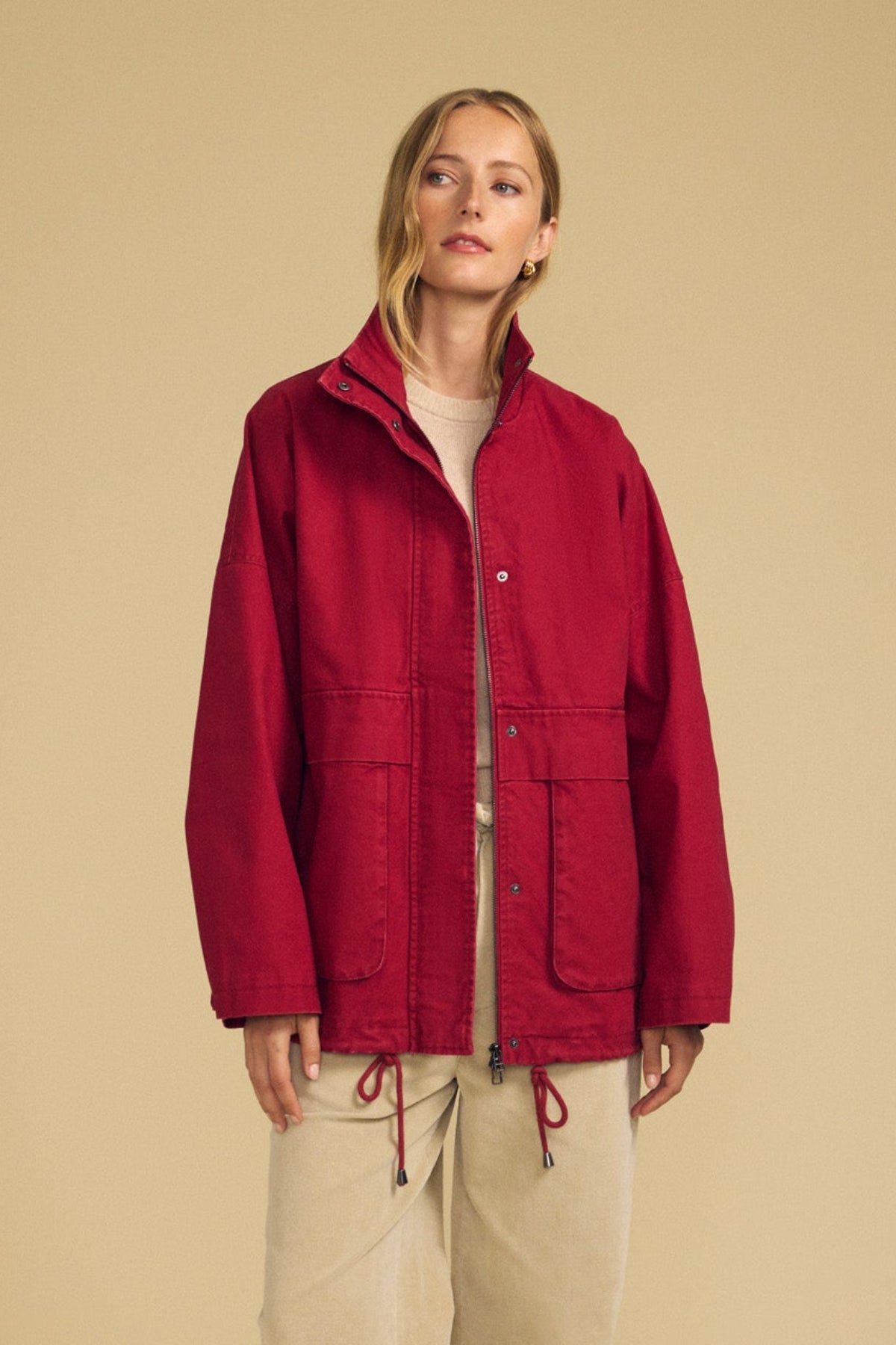 Wearing Velvet by Graham & Spencer’s HUNTER PARKA in red cotton canvas—with oversized front patch pockets—paired with beige pants, a person stands against a plain beige background, ready for cold-weather wear.-41964527452353