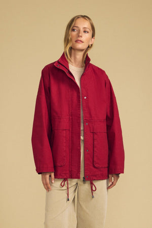 Wearing Velvet by Graham & Spencer’s HUNTER PARKA in red cotton canvas—with oversized front patch pockets—paired with beige pants, a person stands against a plain beige background, ready for cold-weather wear.