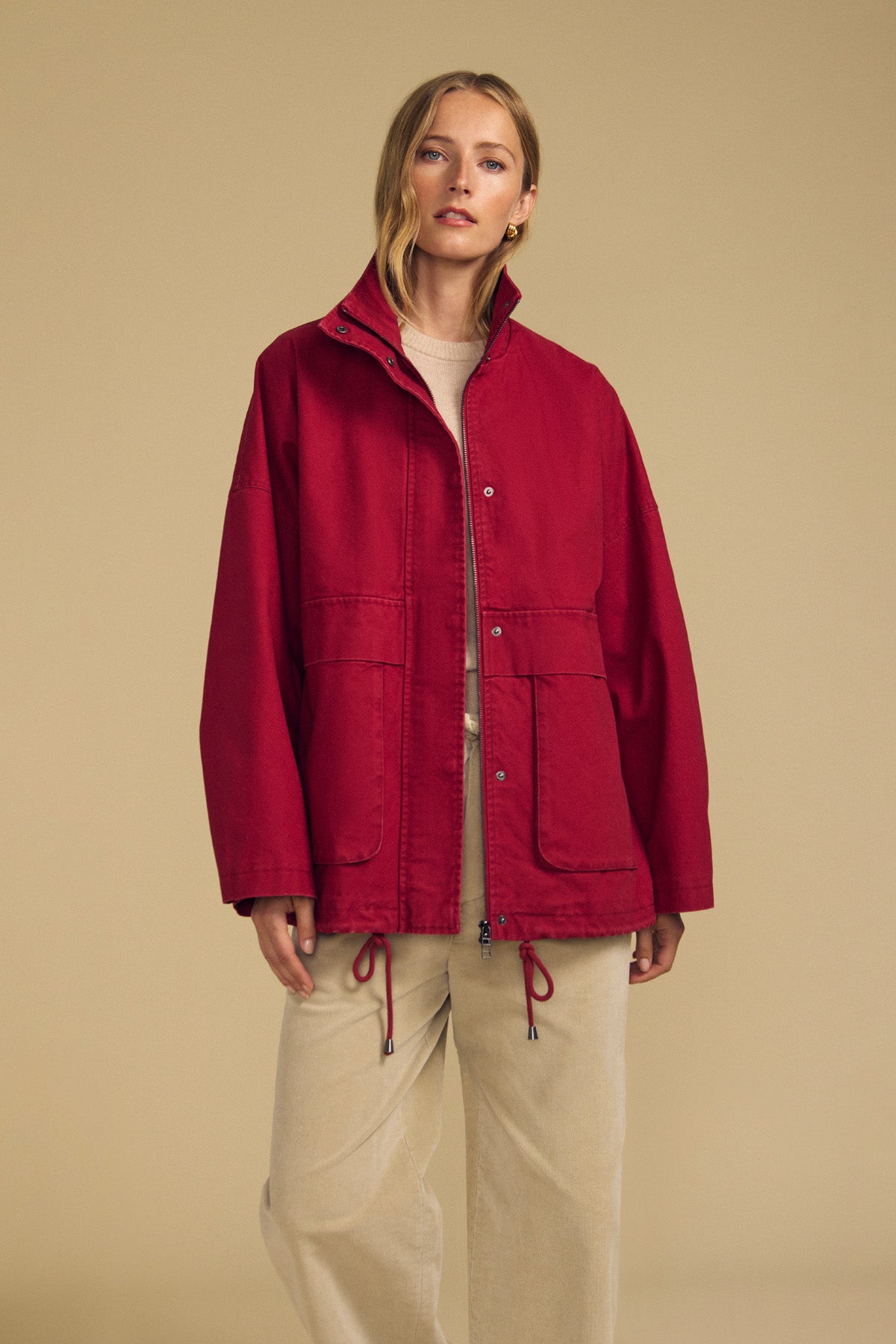 A woman wearing the HUNTER PARKA by Velvet by Graham & Spencer—a loose-fitting red cotton canvas jacket with oversized front patch pockets—layered over a beige top and beige pants, stands against a plain tan background.