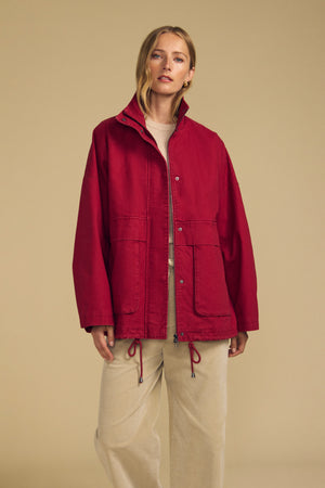 A woman wearing the HUNTER PARKA by Velvet by Graham & Spencer—a loose-fitting red cotton canvas jacket with oversized front patch pockets—layered over a beige top and beige pants, stands against a plain tan background.