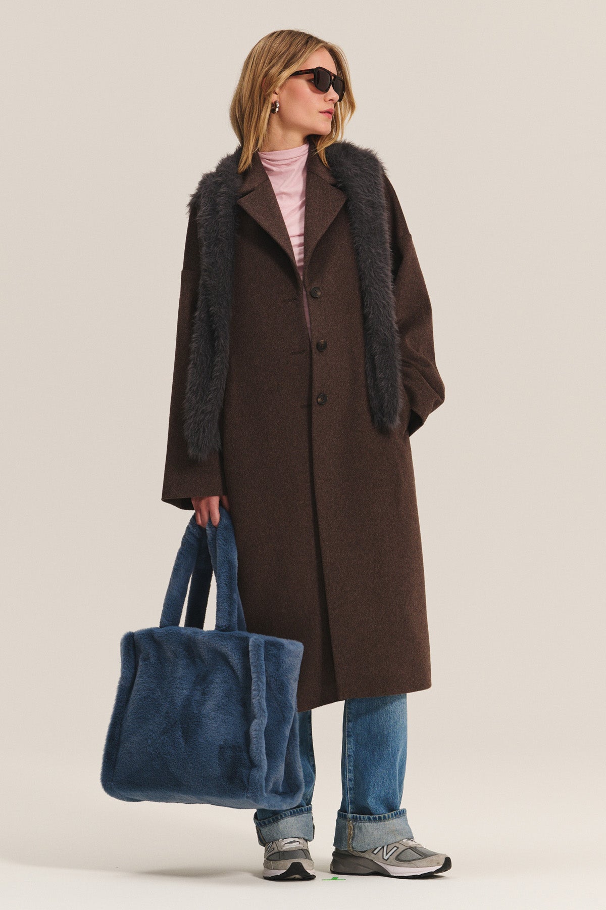A person wearing the LAFAYETTE COAT by Velvet by Graham & Spencer—a long brown wool-blend coat with faux fur trim and classic lapel collar—styled with blue jeans, gray sneakers, sunglasses, and holding a large blue fuzzy tote bag.