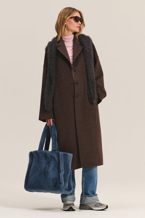 A person wearing the LAFAYETTE COAT by Velvet by Graham & Spencer—a long brown wool-blend coat with faux fur trim and classic lapel collar—styled with blue jeans, gray sneakers, sunglasses, and holding a large blue fuzzy tote bag.