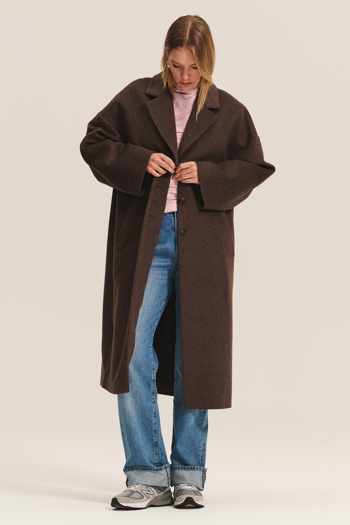 A person fastens the LAFAYETTE COAT by Velvet by Graham & Spencer—a long wool-blend coat with a classic lapel collar—over a pink shirt, blue jeans, and gray sneakers while looking down.-40263652245697