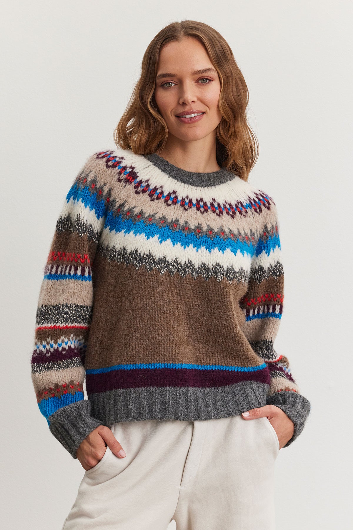   A woman is wearing the KELLA ALPACA FAIR ISLE SWEATER by Velvet by Graham & Spencer, which features a vibrant mix of gray, blue, red, and white designs in a cozy alpaca blend. She pairs it perfectly with light-colored pants. 