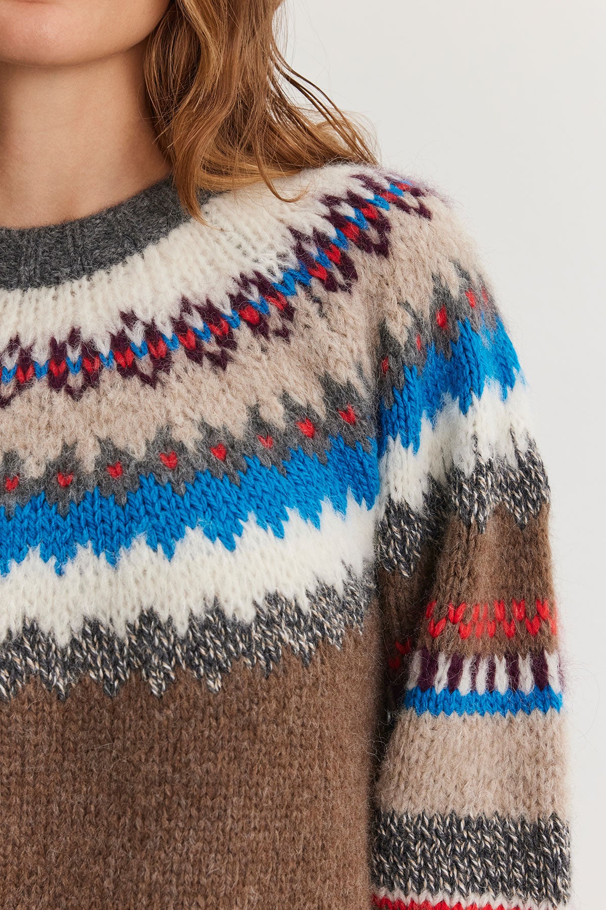   Close-up of a person wearing the KELLA ALPACA FAIR ISLE SWEATER from Velvet by Graham & Spencer, showcasing its colorful geometric pattern in shades of blue, red, white, and brown. Partial view of the person's head and shoulder. 