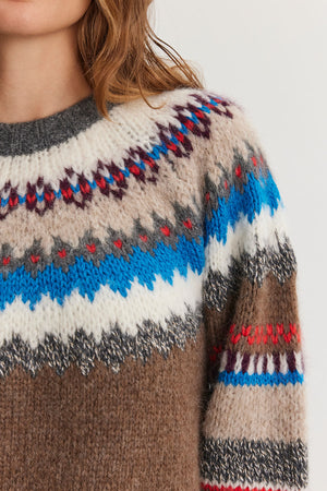 Close-up of a person wearing the KELLA ALPACA FAIR ISLE SWEATER from Velvet by Graham & Spencer, showcasing its colorful geometric pattern in shades of blue, red, white, and brown. Partial view of the person's head and shoulder.