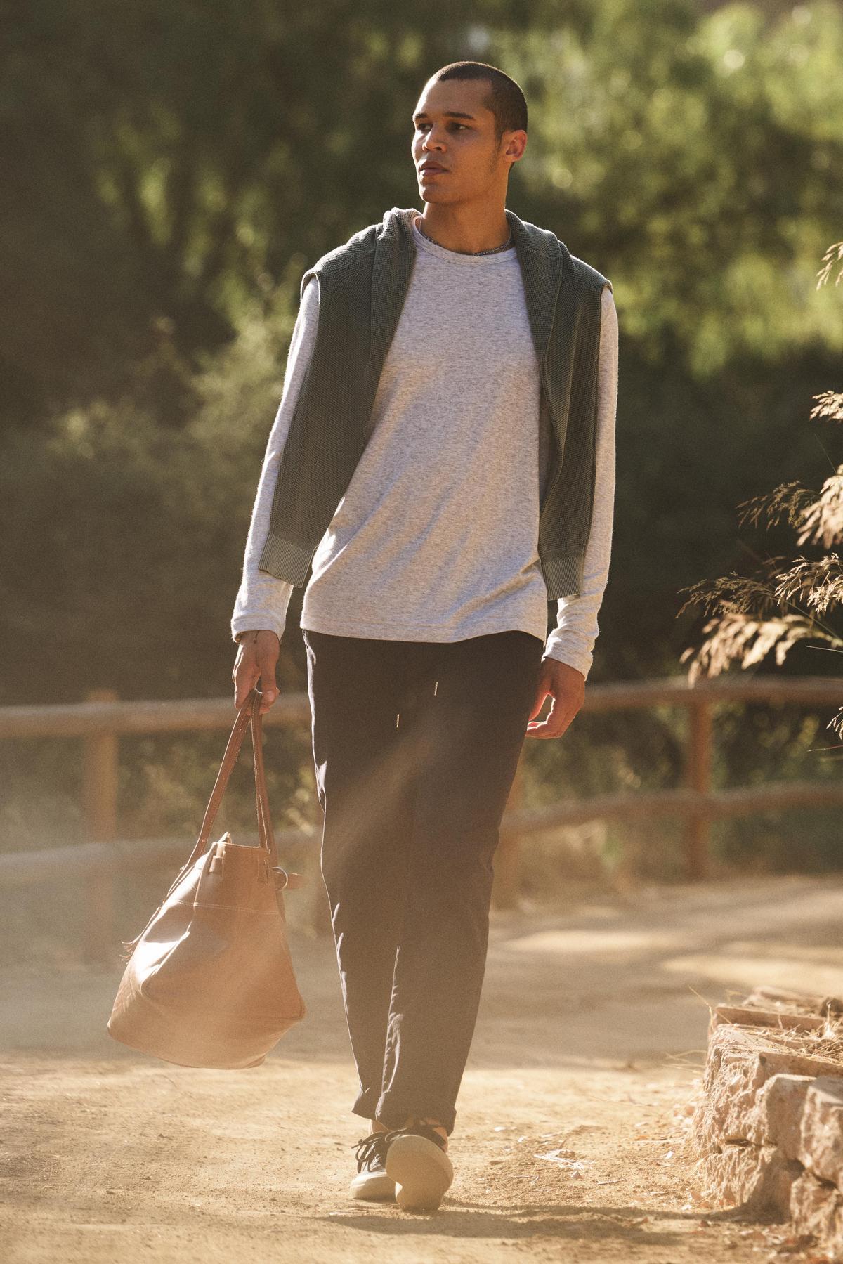 A man walks outdoors on a dirt path wearing the Velvet by Graham & Spencer COLTON CREW, a light long-sleeve with a classic crew neckline, paired with dark pants, a sweatshirt over his shoulders, and carrying a brown bag.