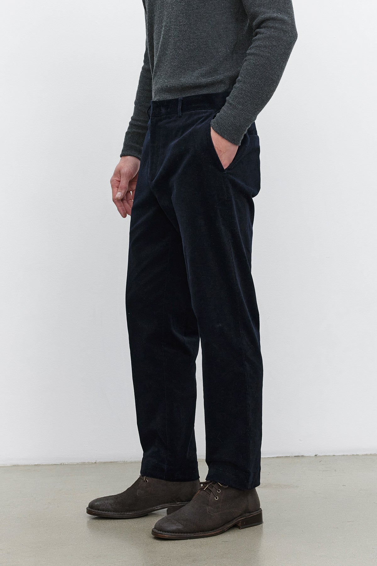   Person standing against a plain background, wearing a gray long-sleeve shirt, Velvet by Graham & Spencer KRYSTOF CORDUROY PANT in dark blue, and brown shoes. Hands are in pockets. 