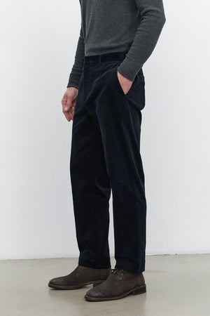 Person standing against a plain background, wearing a gray long-sleeve shirt, Velvet by Graham & Spencer KRYSTOF CORDUROY PANT in dark blue, and brown shoes. Hands are in pockets.