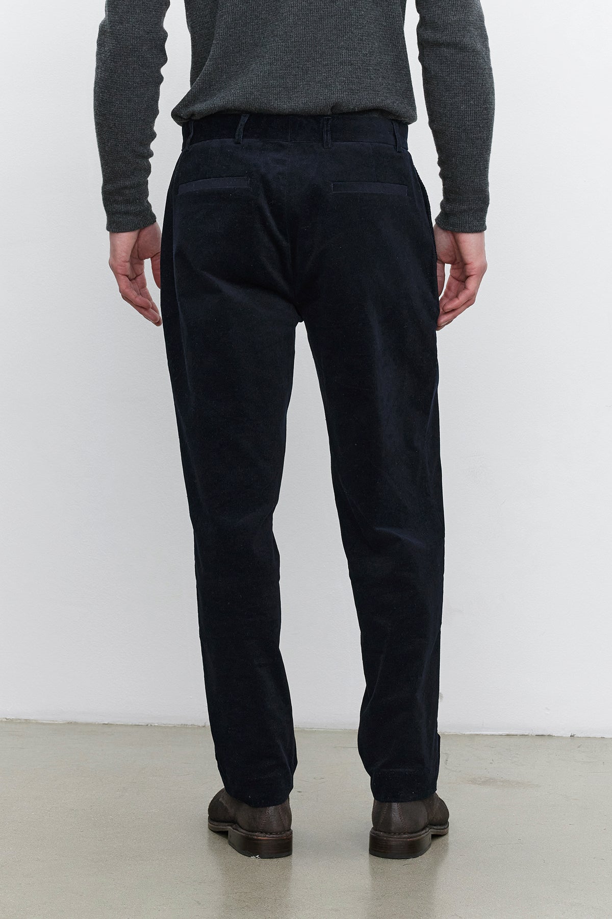   A person standing with their back facing the camera, wearing a dark gray sweater, Velvet by Graham & Spencer's KRYSTOF CORDUROY PANT in dark blue, and brown shoes. 