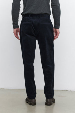 A person standing with their back facing the camera, wearing a dark gray sweater, Velvet by Graham & Spencer's KRYSTOF CORDUROY PANT in dark blue, and brown shoes.