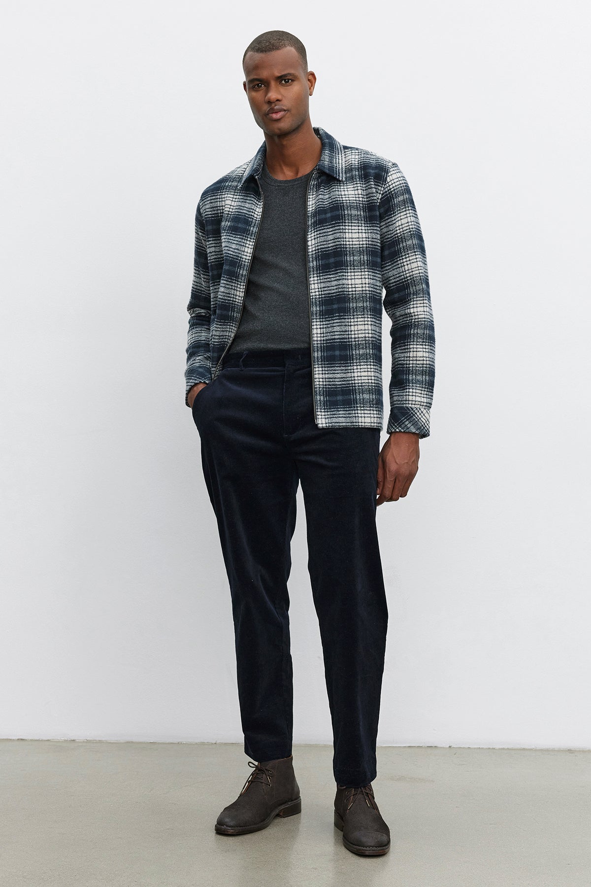   A person stands against a plain background, wearing a gray plaid jacket over a dark sweater, refined and comfortable KRYSTOF CORDUROY PANTS by Velvet by Graham & Spencer tailored in a straight-leg fit, and brown shoes. 