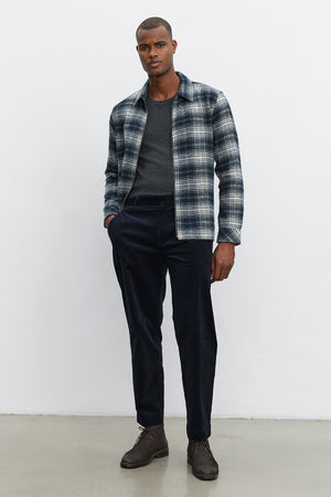 A person stands against a plain background, wearing a gray plaid jacket over a dark sweater, refined and comfortable KRYSTOF CORDUROY PANTS by Velvet by Graham & Spencer tailored in a straight-leg fit, and brown shoes.