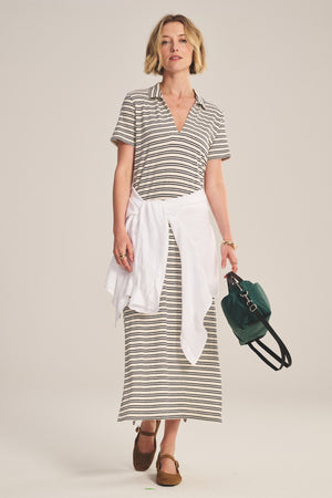 A woman wears the CORIN DRESS by Velvet by Graham & Spencer—a short-sleeve, yarn-dyed stripe jersey maxi—with a white sweater tied at her waist, brown shoes, and a green handbag.
