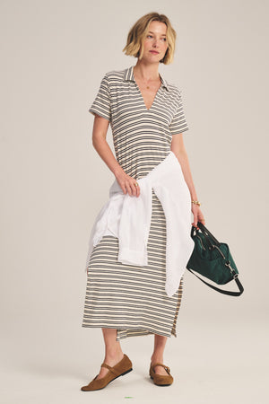 A woman stands against a plain background wearing the CORIN DRESS by Velvet by Graham & Spencer, styled with brown shoes, a white shirt tied at the waist, and holding a green handbag.