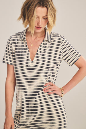 Woman wearing the CORIN DRESS by Velvet by Graham & Spencer, a short-sleeved, black and white striped collared dress, looking down with one hand on her hip against a plain background.