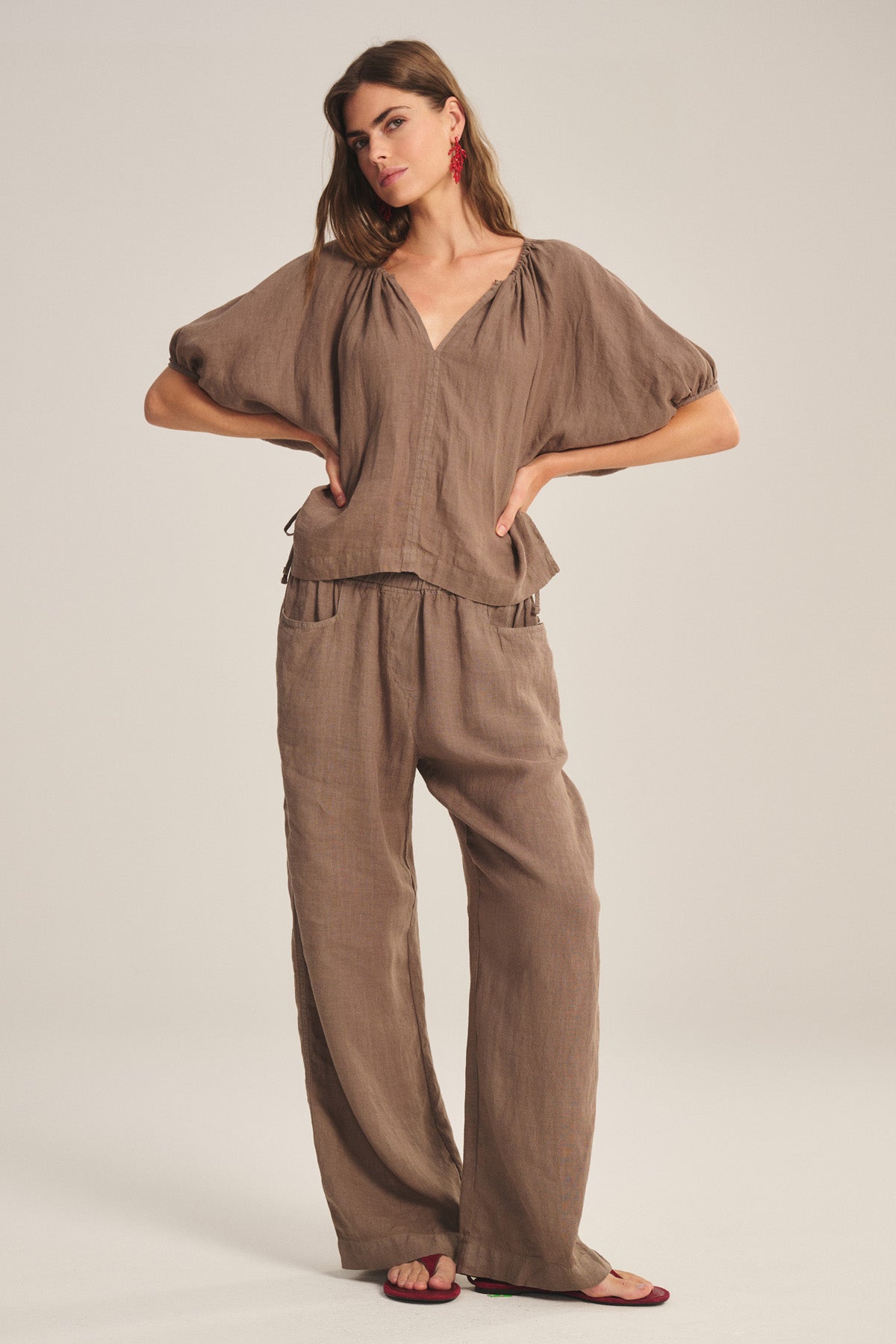 A woman stands in a neutral studio wearing the Velvet by Graham & Spencer CLAIRO LINEN PANT, styled with a loose brown top, red earrings, and sandals.-40909739851969