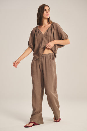 A person stands against a plain background wearing a loose brown short-sleeve top, Velvet by Graham & Spencer’s CLAIRO LINEN PANT with an elastic waist and wide-leg silhouette, and red sandals.