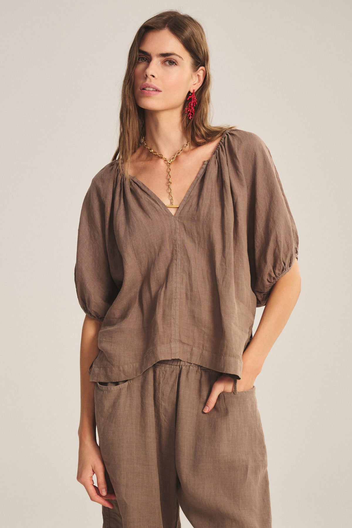A woman wears the Velvet by Graham & Spencer COSETTE LINEN TOP, a relaxed fit, loose brown breathable linen top with short puff sleeves, paired with matching pants, gold necklace, and red earrings.