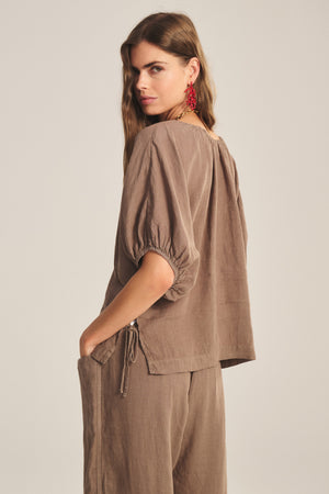 A woman wears the COSETTE LINEN TOP by Velvet by Graham & Spencer, a relaxed-fit brown linen top with elbow-length sleeves and matching pants, standing with one hand in her pocket and glancing over her shoulder.
