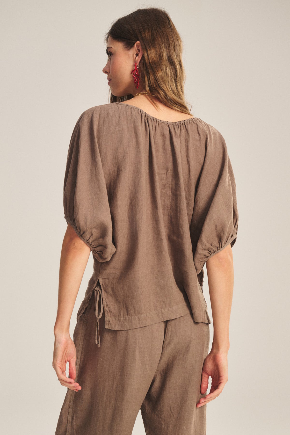 A woman is shown from the back wearing the COSETTE LINEN TOP by Velvet by Graham & Spencer—a relaxed fit, breathable linen piece with puffed sleeves—paired with matching brown pants.-40909829439681