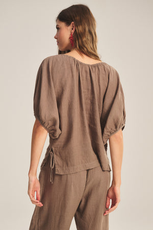 A woman is shown from the back wearing the COSETTE LINEN TOP by Velvet by Graham & Spencer—a relaxed fit, breathable linen piece with puffed sleeves—paired with matching brown pants.