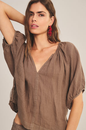A woman wears the COSETTE LINEN TOP by Velvet by Graham & Spencer, a relaxed fit brown blouse with a deep V neckline and short sleeves, paired with red dangling earrings against a neutral background.