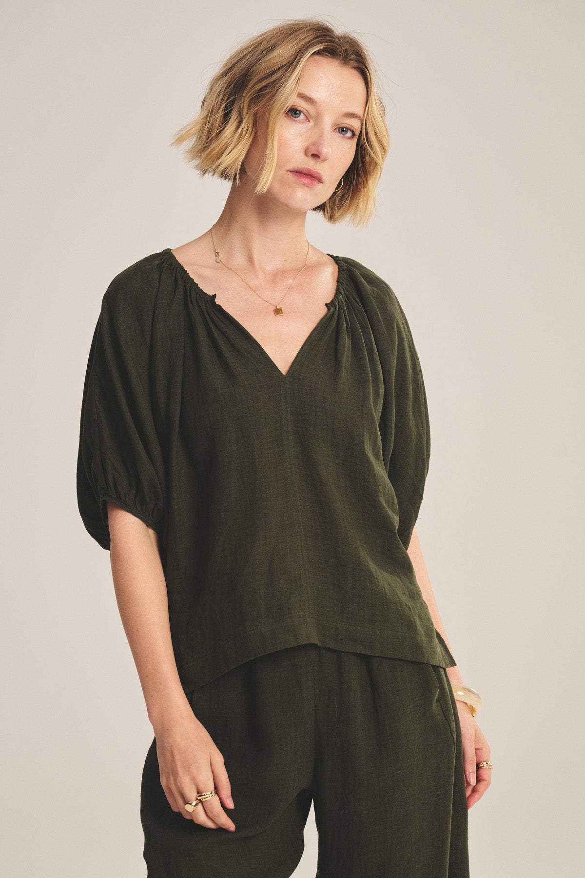A woman with short blonde hair wears the COSETTE LINEN TOP by Velvet by Graham & Spencer—an olive green, woven linen top with a deep V neckline and relaxed fit—paired with matching pants, against a plain light background.-40808147288257