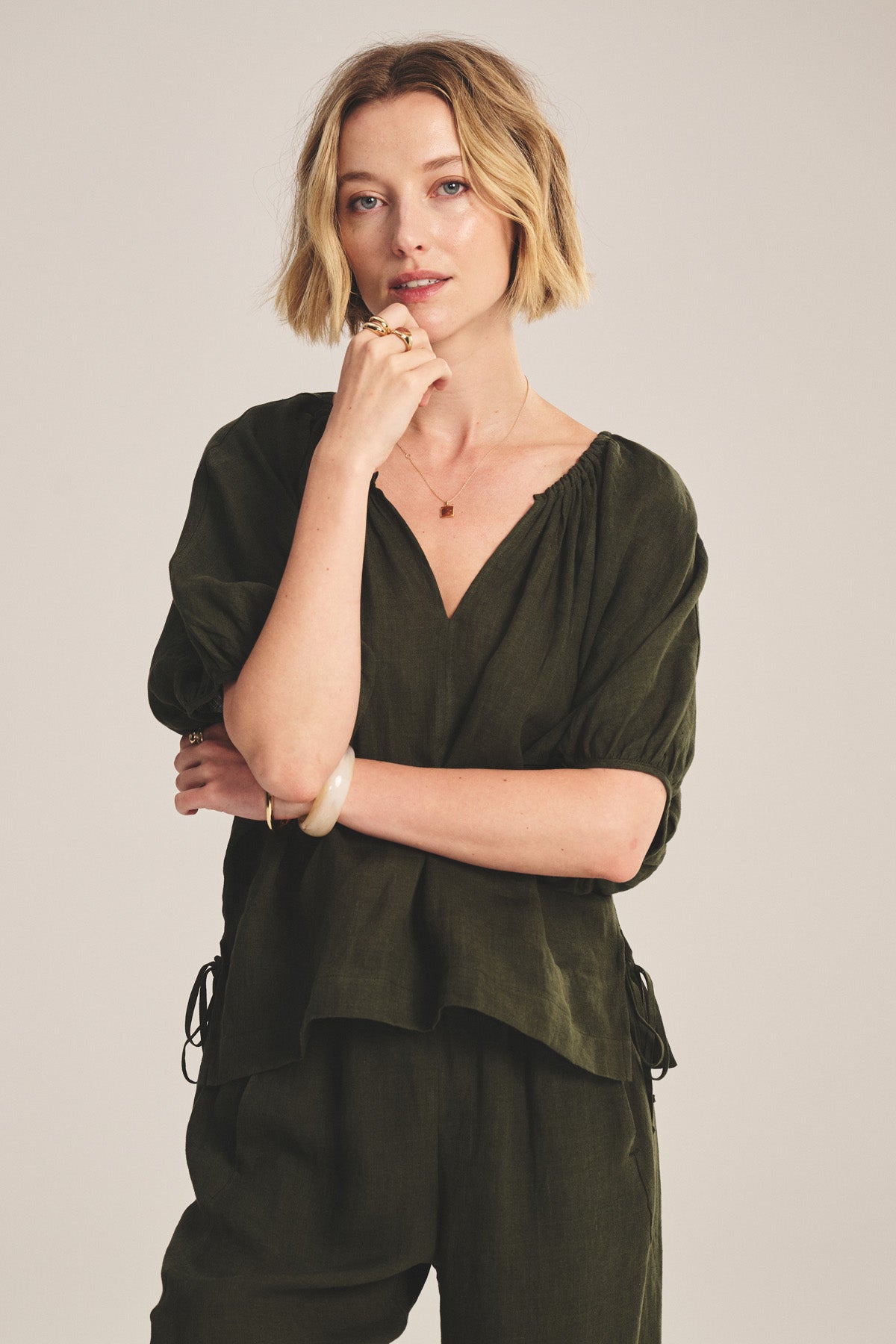 A woman with short blonde hair models the COSETTE LINEN TOP in olive green by Velvet by Graham & Spencer, paired with matching pants, posing with one hand on her chin against a plain background.