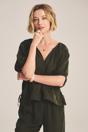 A woman with short blonde hair models the COSETTE LINEN TOP in olive green by Velvet by Graham & Spencer, paired with matching pants, posing with one hand on her chin against a plain background.