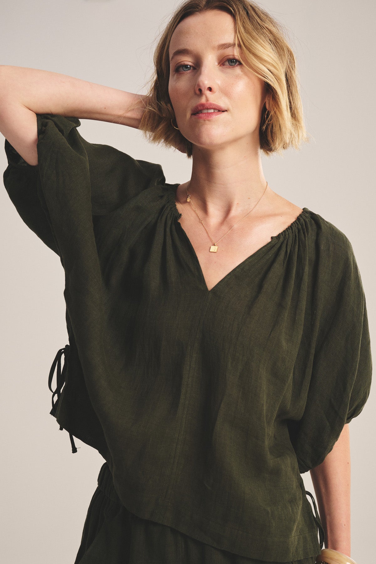 A woman with short blonde hair models the COSETTE LINEN TOP by Velvet by Graham & Spencer, a relaxed fit olive green linen blouse with a deep V neckline, paired with a gold necklace and standing with one hand behind her head.