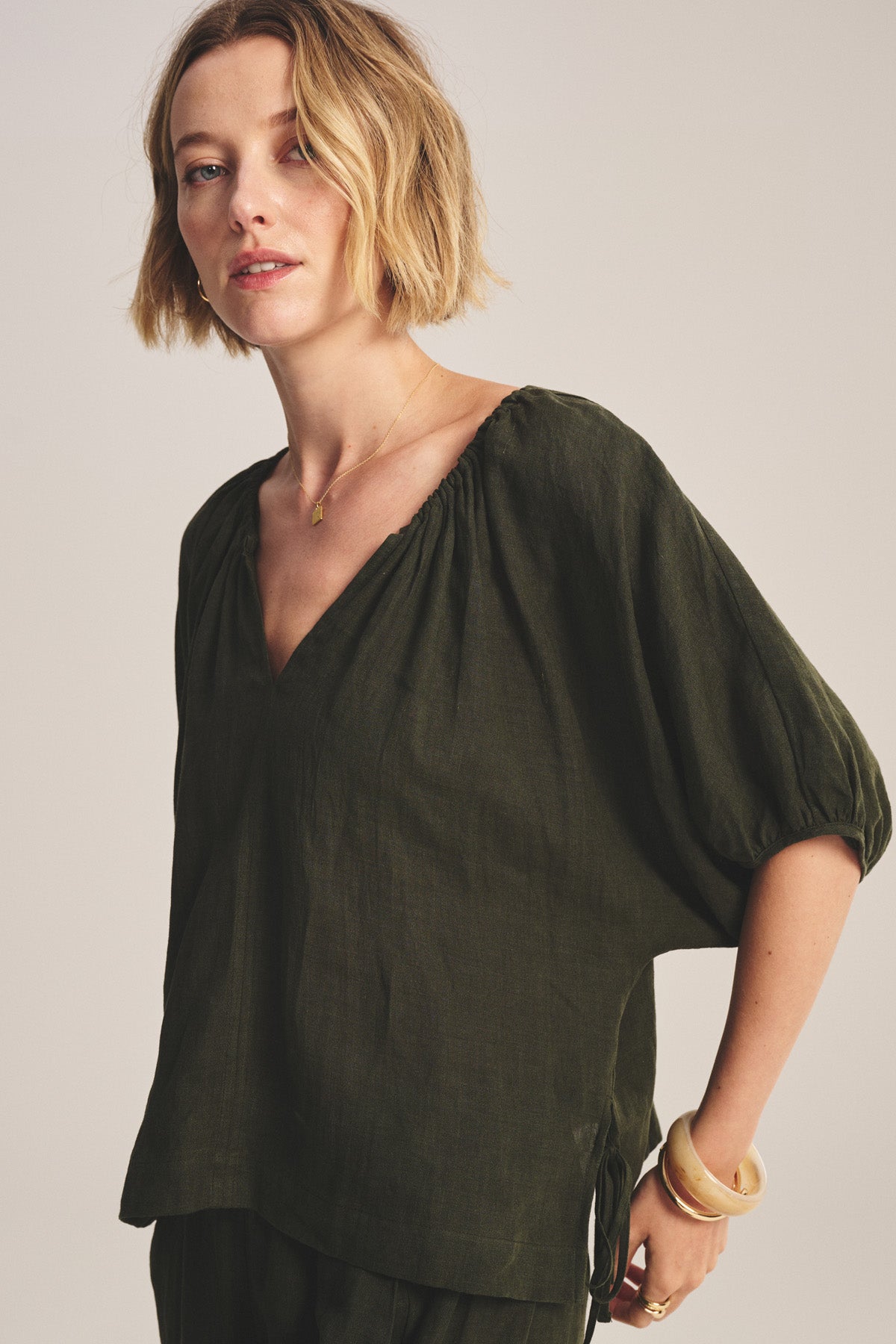 A woman with short blonde hair wears the COSETTE LINEN TOP by Velvet by Graham & Spencer, a dark green breathable linen top with a deep V neckline and gold jewelry, standing against a neutral background.-40808147452097