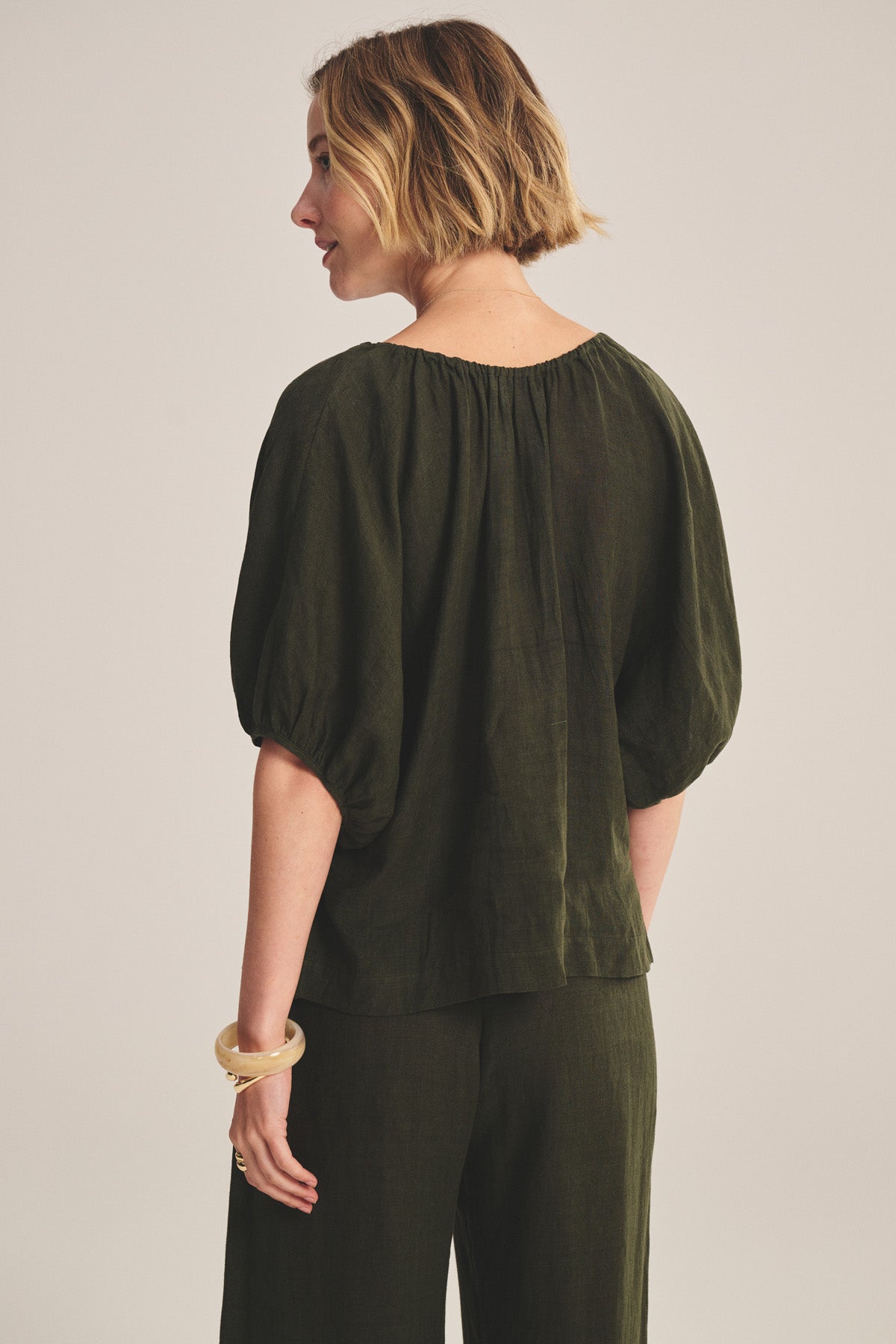 A woman with short, wavy hair faces away, wearing Velvet by Graham & Spencer's COSETTE LINEN TOP—a relaxed dark green woven linen piece with elbow-length sleeves and matching pants, styled with a chunky bracelet.-40808147517633