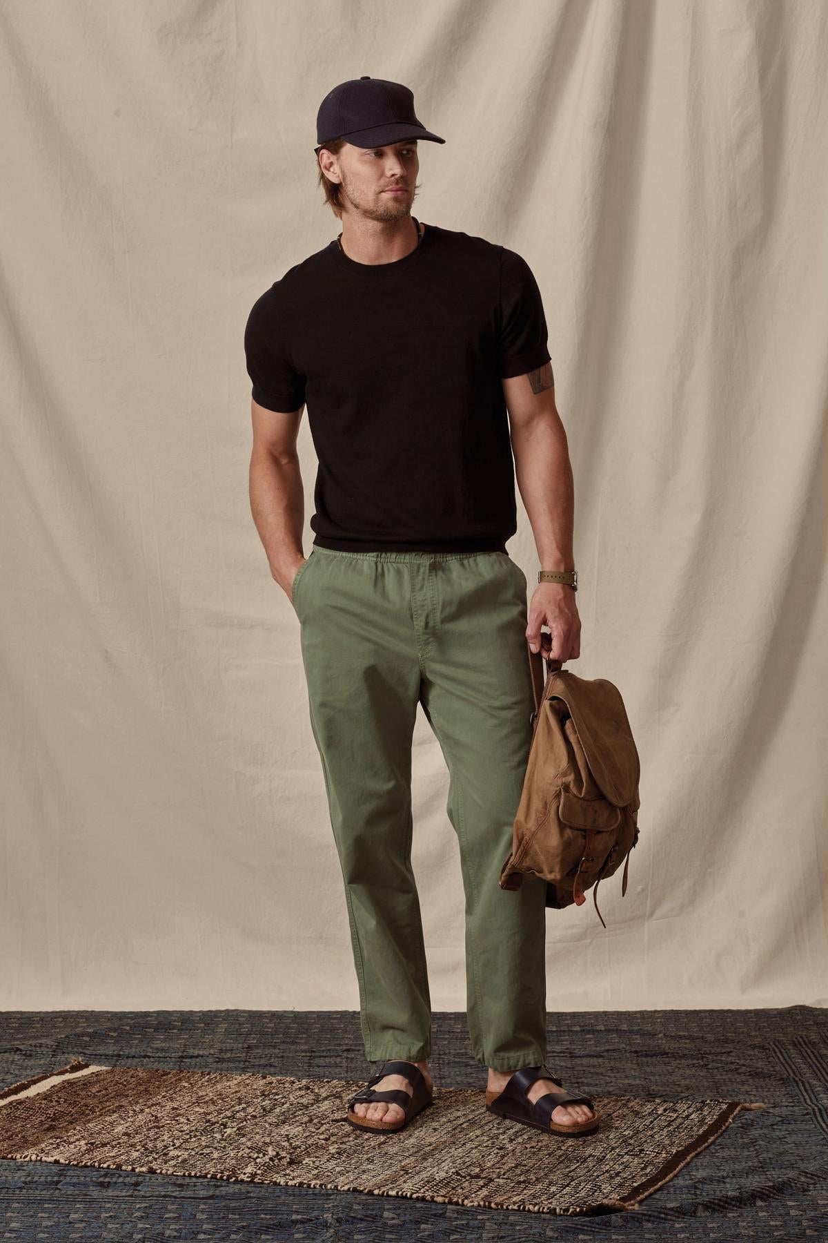   Man in a casual black t-shirt and green Branson Pant by Velvet by Graham & Spencer with an elastic drawstring waist holding a backpack, wearing a cap and sandals, standing against a draped backdrop. 