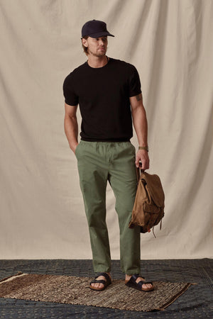 Man in a casual black t-shirt and green Branson Pant by Velvet by Graham & Spencer with an elastic drawstring waist holding a backpack, wearing a cap and sandals, standing against a draped backdrop.