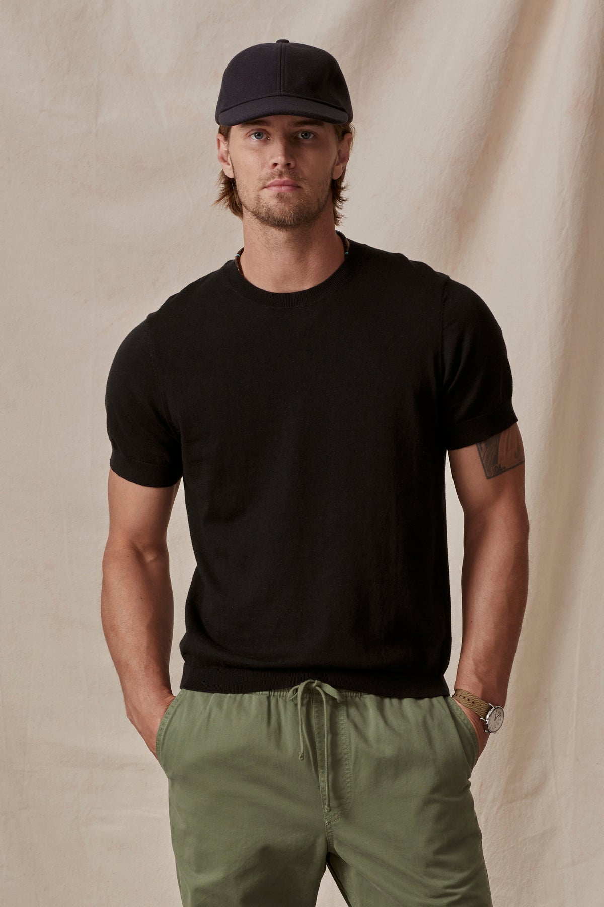 A man wearing a black cotton/linen blend Dexter Crew t-shirt from Velvet by Graham & Spencer and green trousers, accessorized with a cap and a watch, poses against a beige background.-36753529307329