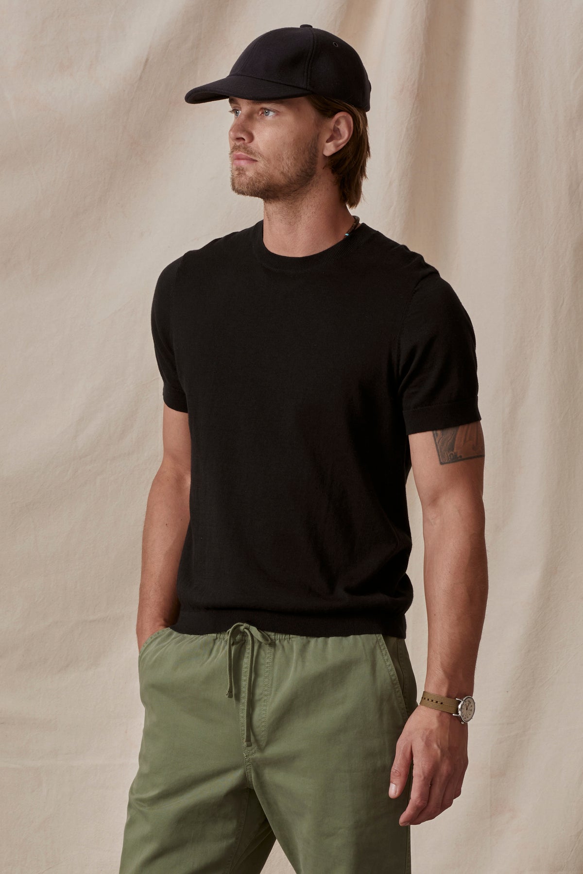 A man in a Velvet by Graham & Spencer cotton/linen blend black Dexter Crew t-shirt and green pants with a black cap, standing against a beige background, looking to the side.-36753529340097