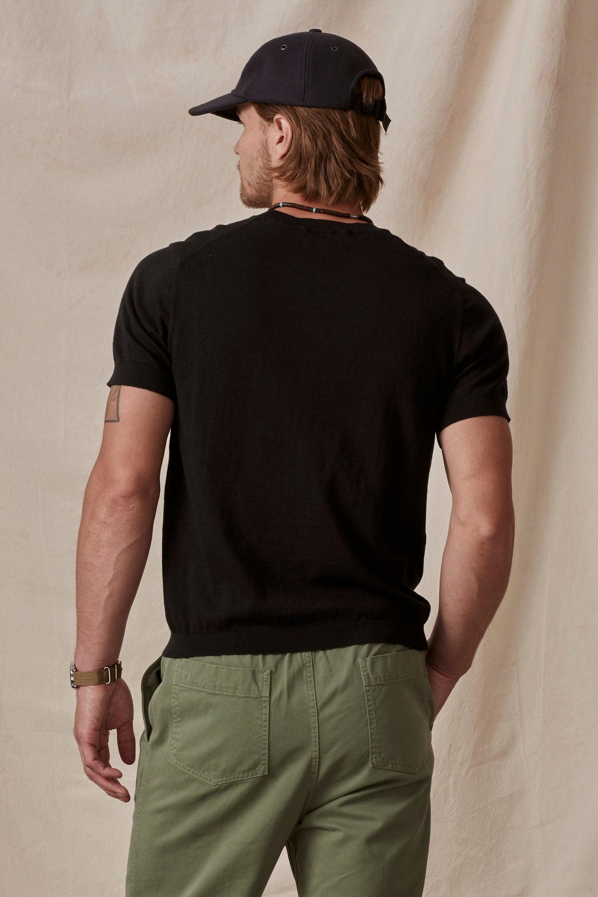 A man viewed from the back, wearing a black cotton/linen blend t-shirt (DEXTER CREW by Velvet by Graham & Spencer) and green pants, with a baseball cap, standing against a beige backdrop.-36753529372865