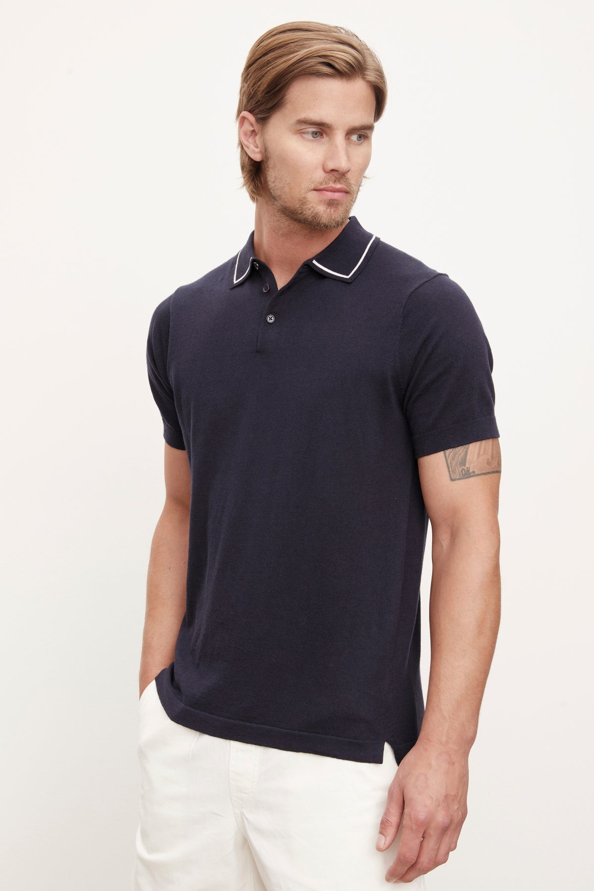 A man in a Velvet by Graham & Spencer Shepard polo shirt and white pants standing against a light background, looking to the side with a subtle expression.-36753534746817