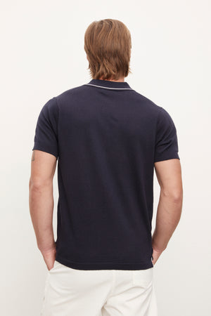 Man wearing a Velvet by Graham & Spencer Shepard Polo shirt in navy blue and white pants, standing with his back to the camera, against a white background.