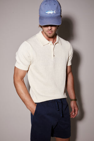 Man wearing a TIBERIUS POLO by Velvet by Graham & Spencer polo shirt, navy shorts, and a blue cap with a shark emblem, standing against a light brown background.