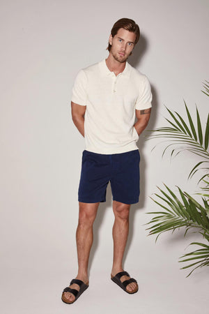 A man in a white polo shirt and navy blue Velvet by Graham & Spencer FIELDER SHORT stands confidently against a light background with a potted plant nearby.