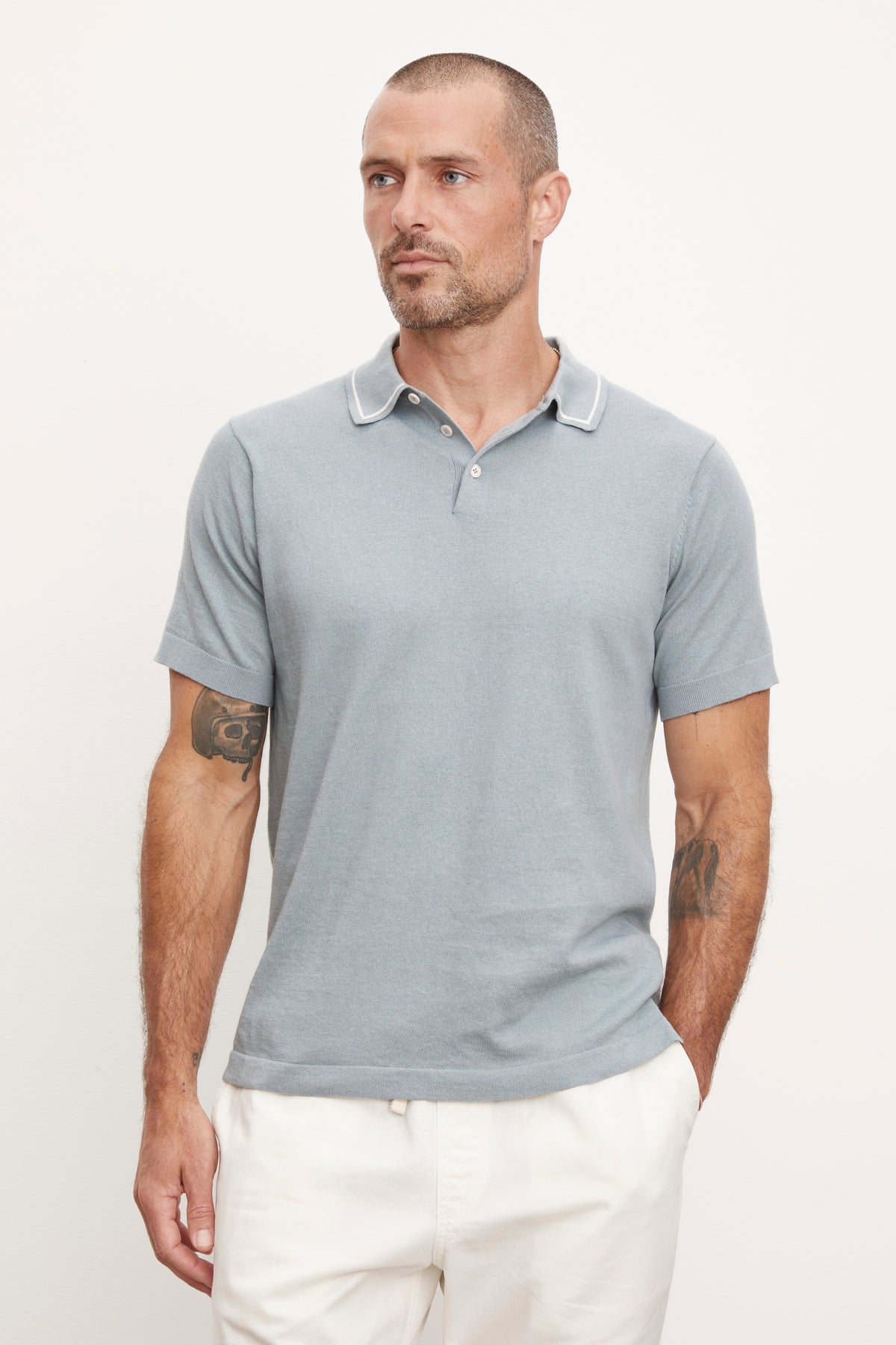   Man in a Velvet by Graham & Spencer Shepard Polo shirt and white pants, looking to the side with visible tattoos on both arms. 