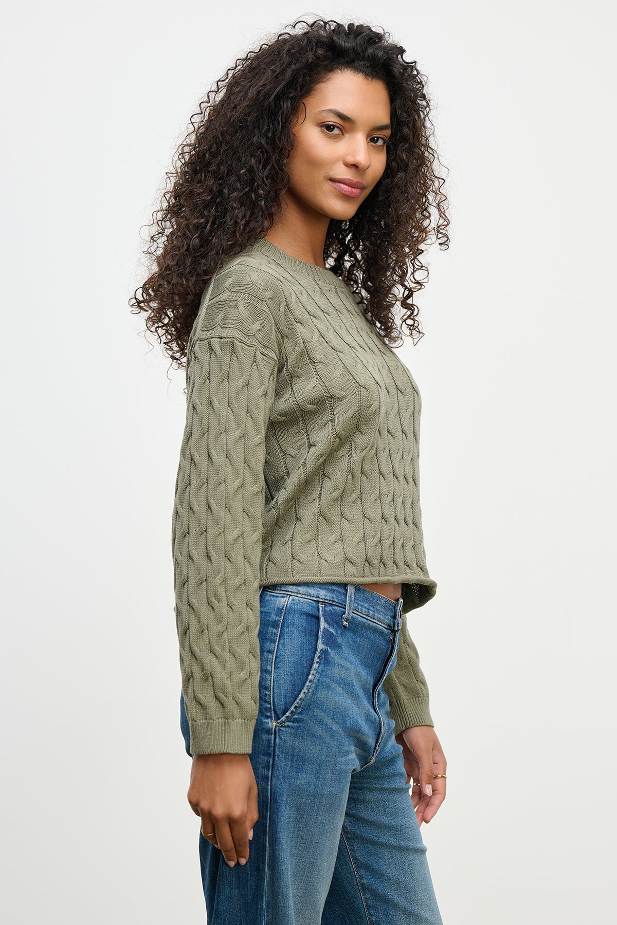 A person with curly hair is wearing a green APRIL SWEATER by Velvet by Graham & Spencer and blue jeans, standing against a plain background.-38662829113537